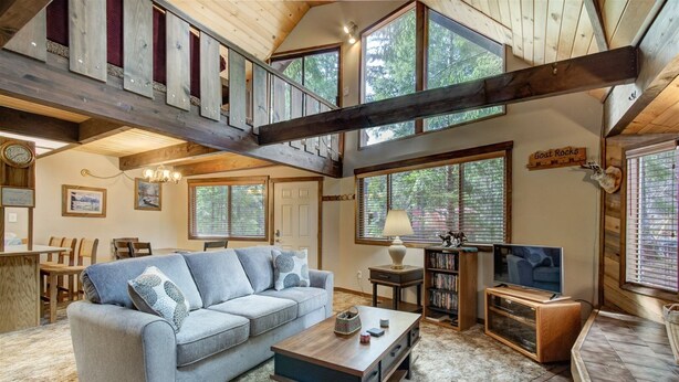 A spacious living room with a grey sofa, a coffee table, and a TV. There is a staircase with wooden beams and a large window with blinds. - Lake Creek Cabin (Packwood)
