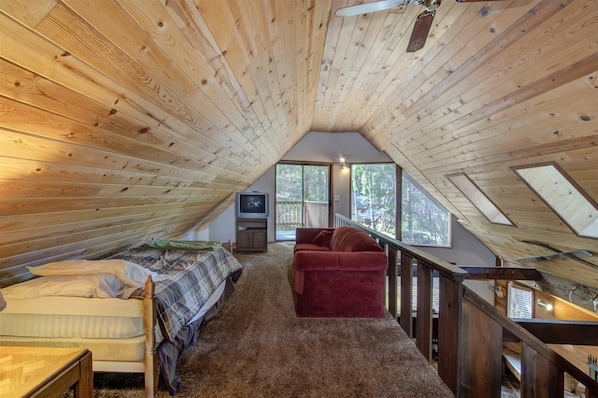 An attic bedroom with a sloped ceiling, a bed, a red sofa, a TV, and a view of the outdoors.