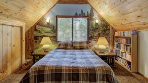 A cozy attic bedroom with a large window, wooden walls, a bed with plaid bedding, two bedside lamps, and a bookshelf. - Lake Creek Cabin (Packwood)