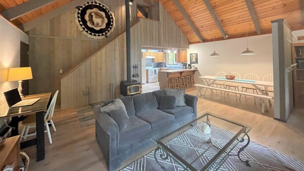 A cozy living room with a gray sofa, a glass coffee table, and a wooden ceiling. A kitchen is visible in the background with wooden cabinets and a dining area with white chairs. - Rainier Retreat (Packwood)