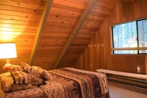 A cozy attic bedroom with a wooden ceiling, a large window, a bed with patterned bedding, and a bedside table.
