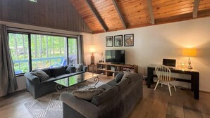 A spacious living room with a large sofa, a coffee table, and a TV. There are three framed pictures on the wall, a desk with a chair, and a window with a view of trees. - Rainier Retreat (Packwood)
