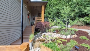 A house with a wooden front porch, a stone retaining wall, and a garden with various plants and a sign that says "LOOSE ROCK Please stay off." - Ravenwood (Packwood)