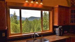 A kitchen with a large window offering a view of a forest and mountains. - Ravenwood (Packwood)
