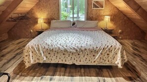 An attic bedroom with a sloped ceiling, a large bed with floral bedding, two bedside lamps, and a window with a view of greenery.