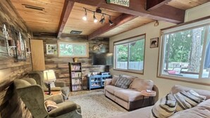 A cozy living room with a fireplace, a sofa, a chair, and a bookshelf. There is a large window with a view of trees.