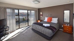 A hotel room with a large bed, a television, a dresser, and a balcony with a view of buildings. - ASURE Adrian Motel (Dunedin)