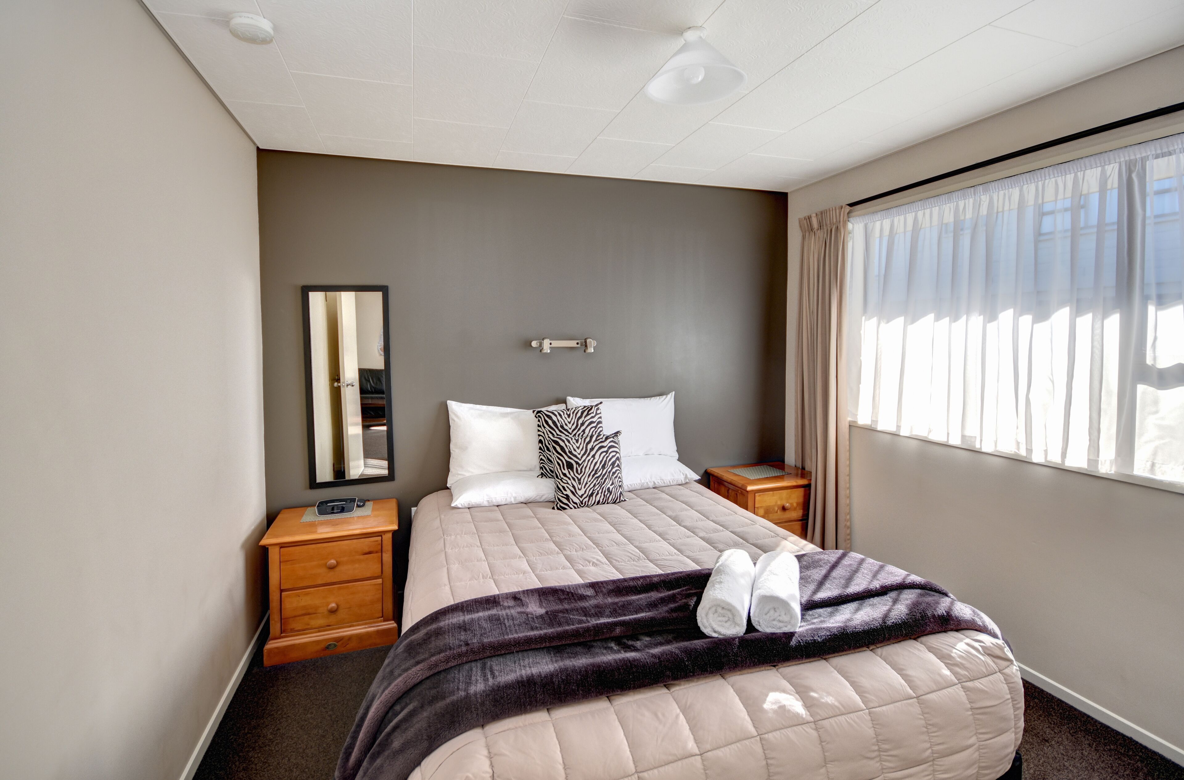 A hotel room with a bed, a nightstand, a bedside table, a mirror, and a window with curtains.