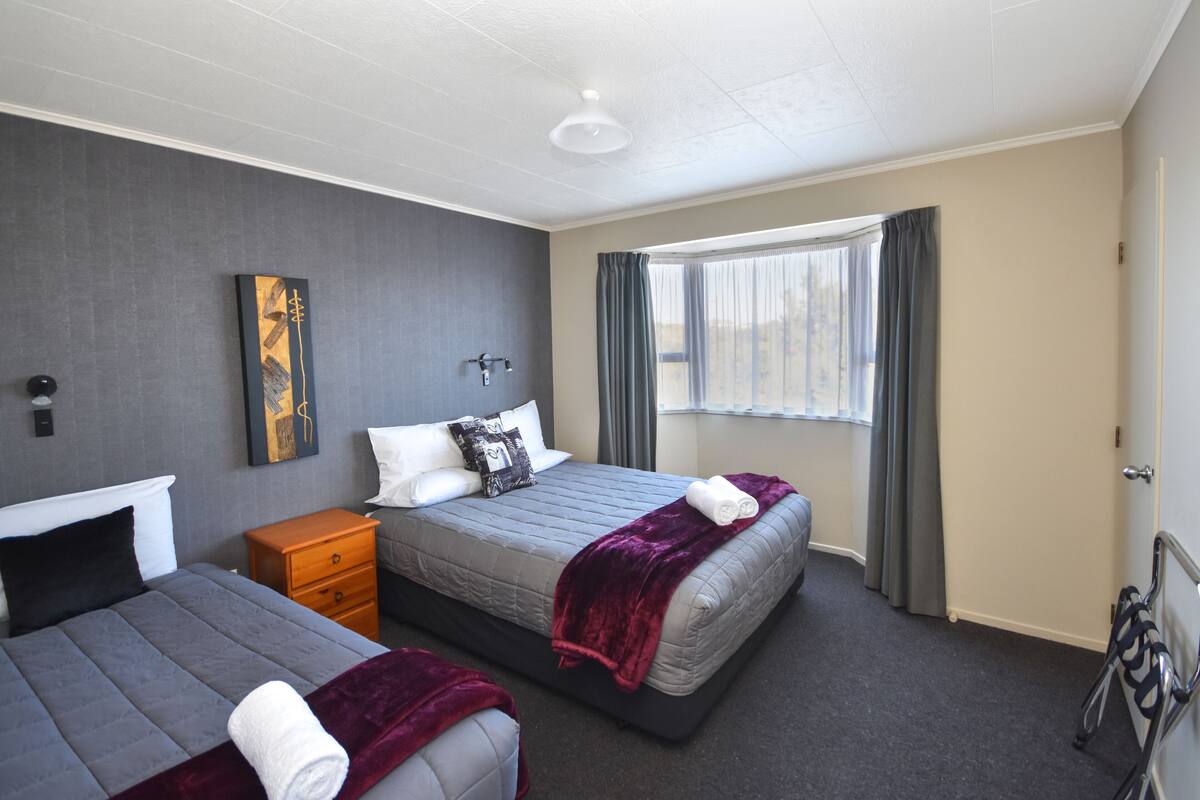 A hotel room with two beds, a nightstand, a painting on the wall, a window with curtains, and a ceiling light.