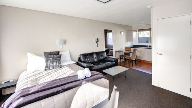 A hotel room with a bed, a black leather sofa, a small table, and a kitchenette in the background. - ASURE Adrian Motel (Dunedin)