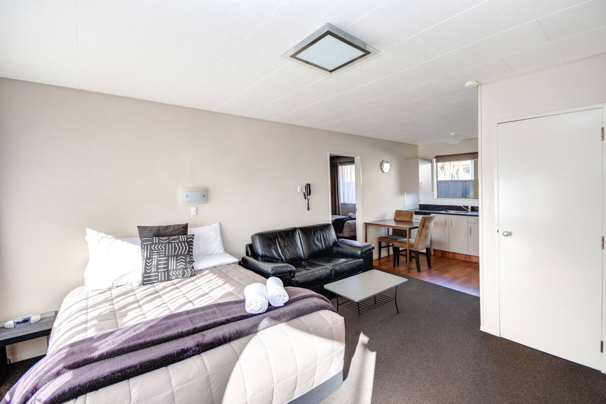 A hotel room with a bed, a black leather sofa, a small table, and a kitchenette in the background.