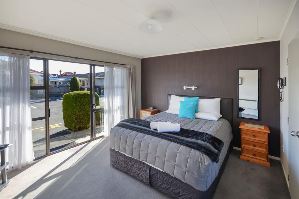 A modern hotel room with a large bed, a dresser, a mirror, and a sliding glass door leading to a balcony.
