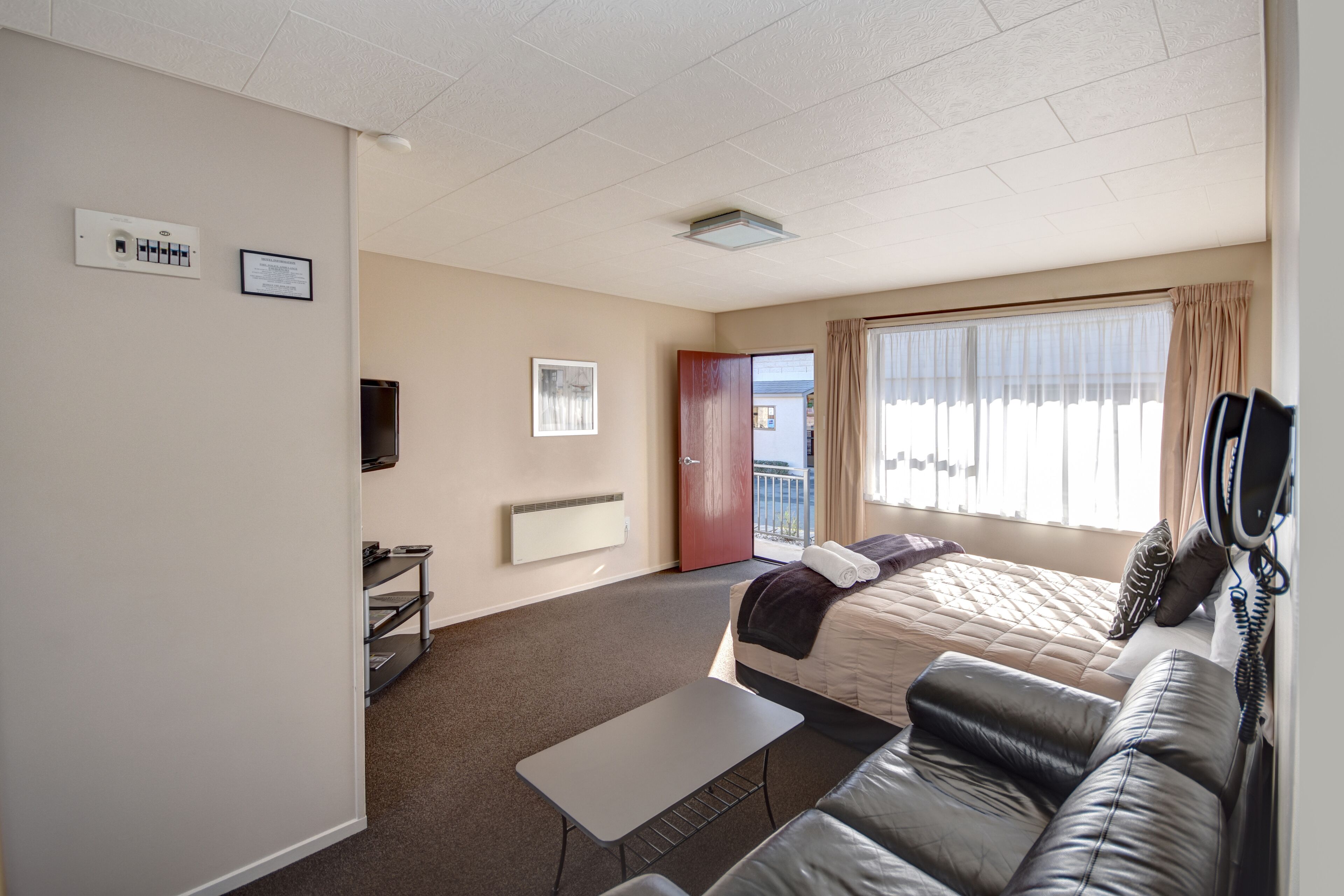 A hotel room with a bed, a small table, a sofa, a radiator, a window with curtains, a door, and a wall-mounted phone.