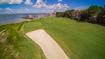 Un campo da golf con un faro sullo sfondo, una distesa d'acqua e una serie di edifici.