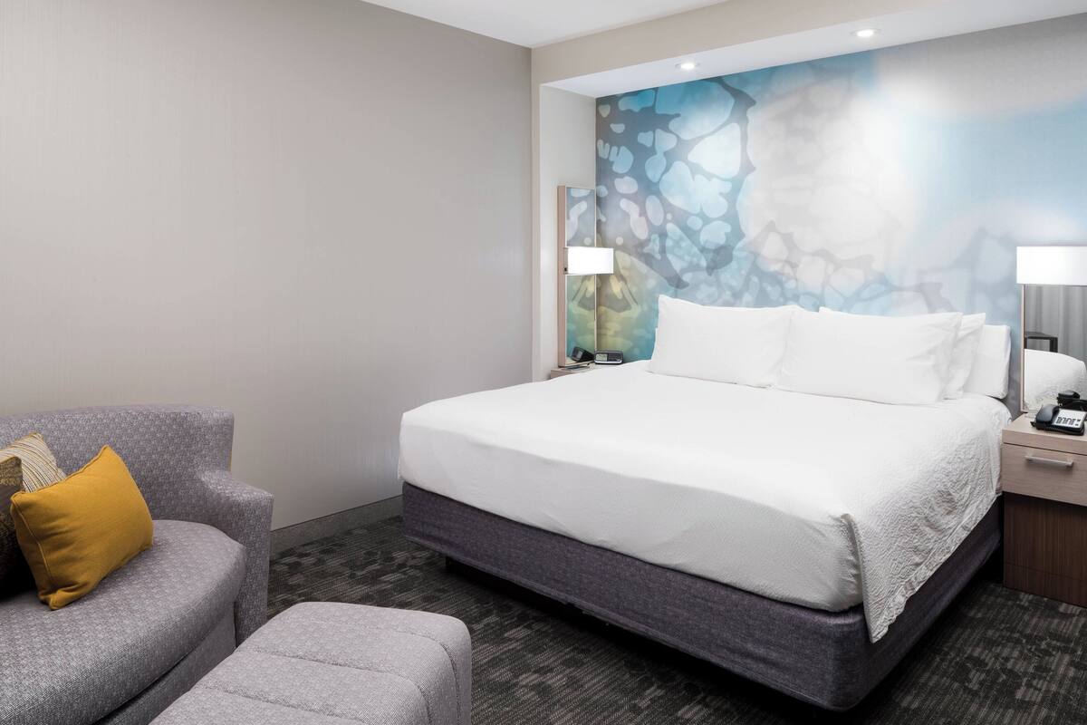 Een moderne hotelkamer met een groot bed, een grijze fauteuil, een klein poufje, en een muur met een blauw abstract ontwerp.