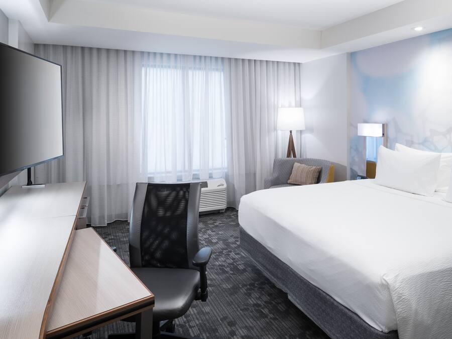 A hotel room with a large bed, a desk with a chair, a flat-screen TV, and a large window with curtains.