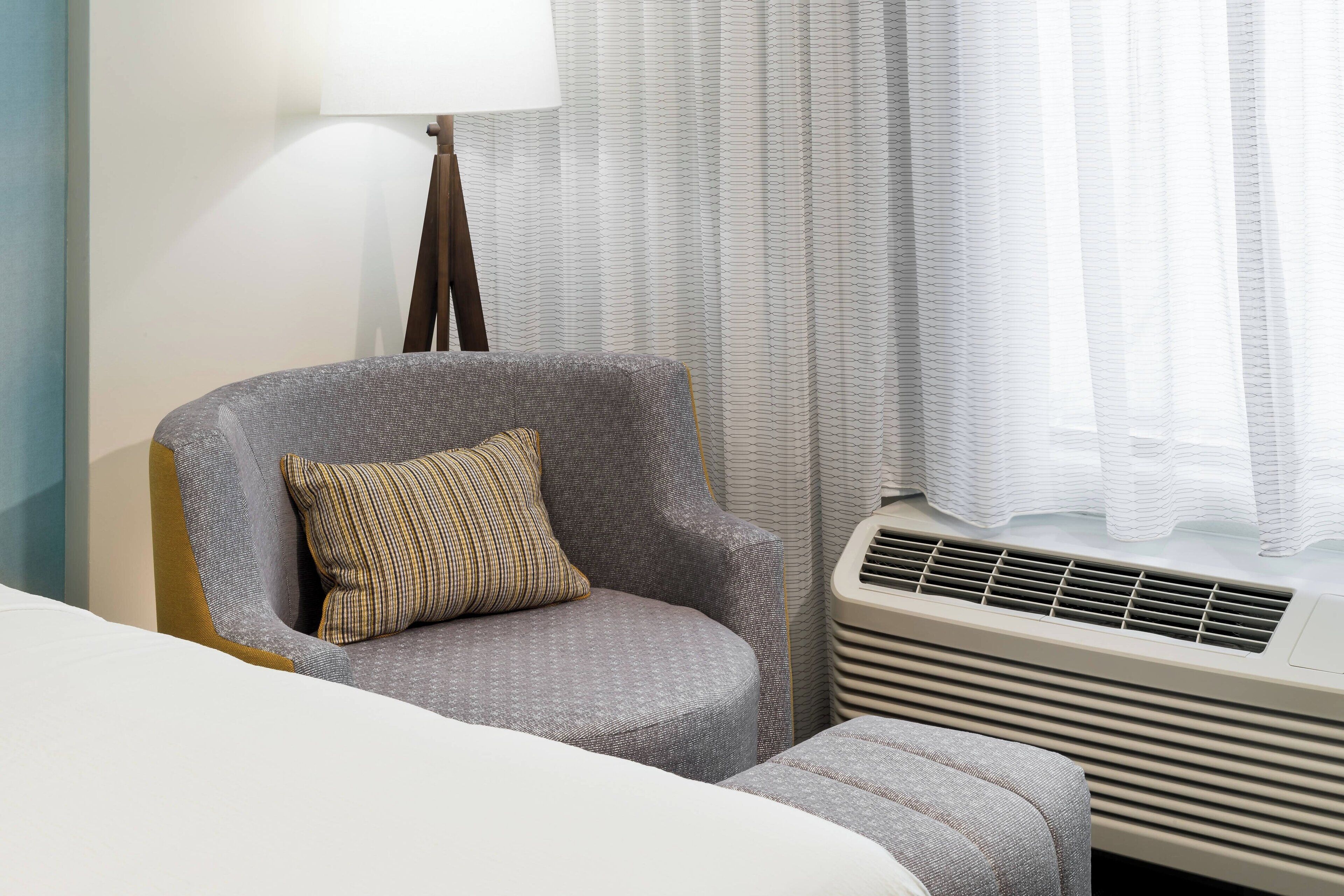 Una habitación de hotel con un sillón gris, un pequeño reposapiés, una cama blanca, una lámpara de pie y una ventana con cortinas traslúcidas.