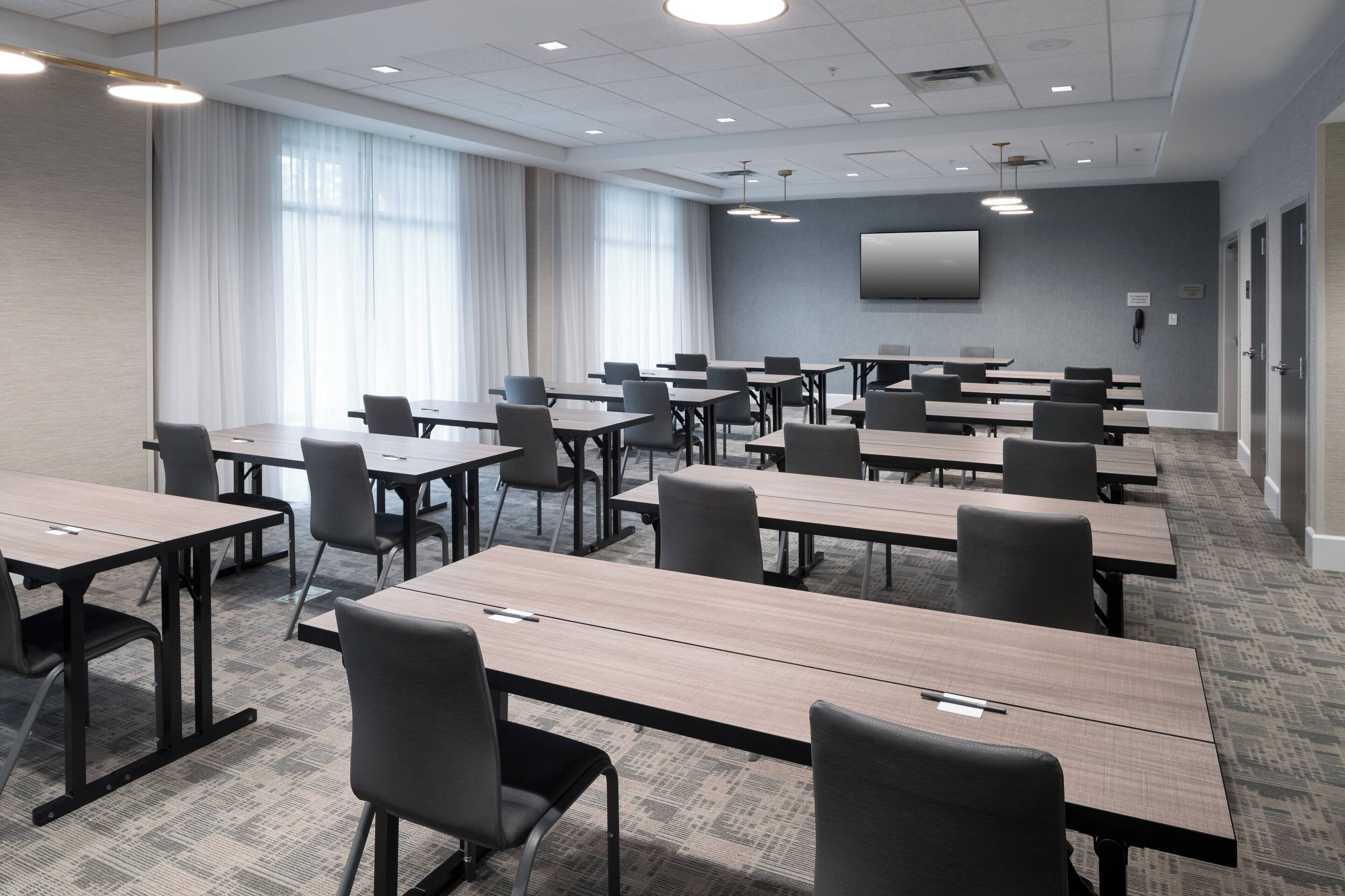 A conference room with multiple tables and chairs, large windows with curtains, a flat-screen TV on the wall, and a carpeted floor.