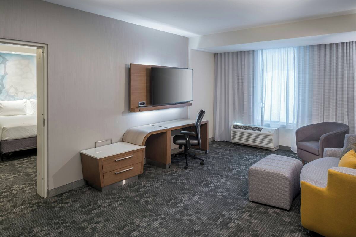 Een hotelkamer met een bed, een bureau met stoel, een flatscreen televisie boven het bureau, een groot raam met gordijnen en een zithoek met een bank en voetenbankje.