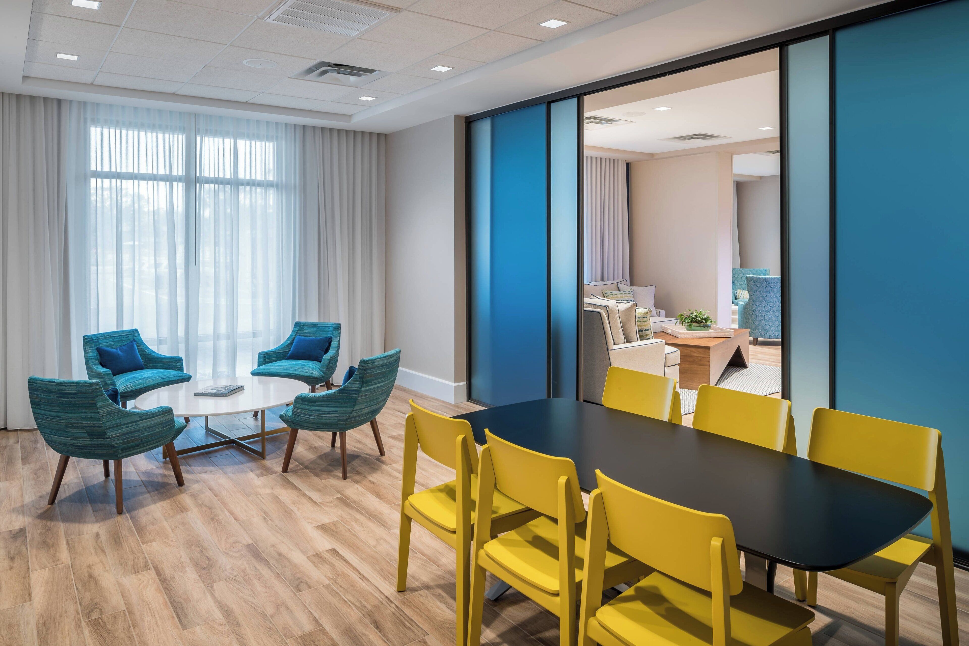Una sala de reuniones moderna con una mesa grande y sillas amarillas, sillas azules, y vista a un área de dormitorio.
