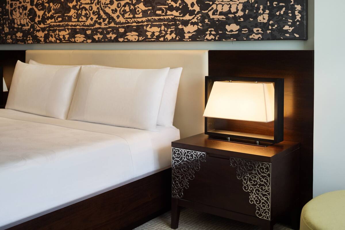Una cama bien hecha con sábanas blancas, una lámpara de noche y un adorno decorativo en la pared.
