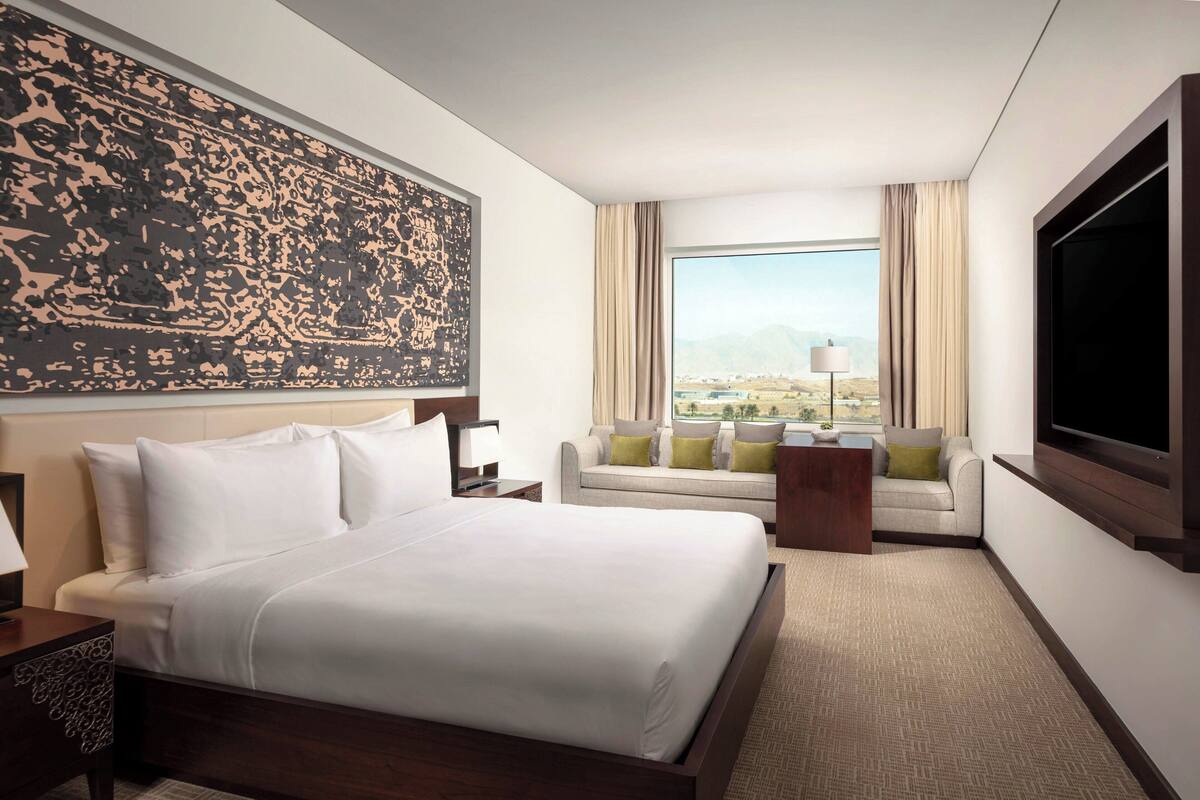 Habitación de hotel con una cama grande, un sofá, un televisor y una ventana con vistas a las montañas.