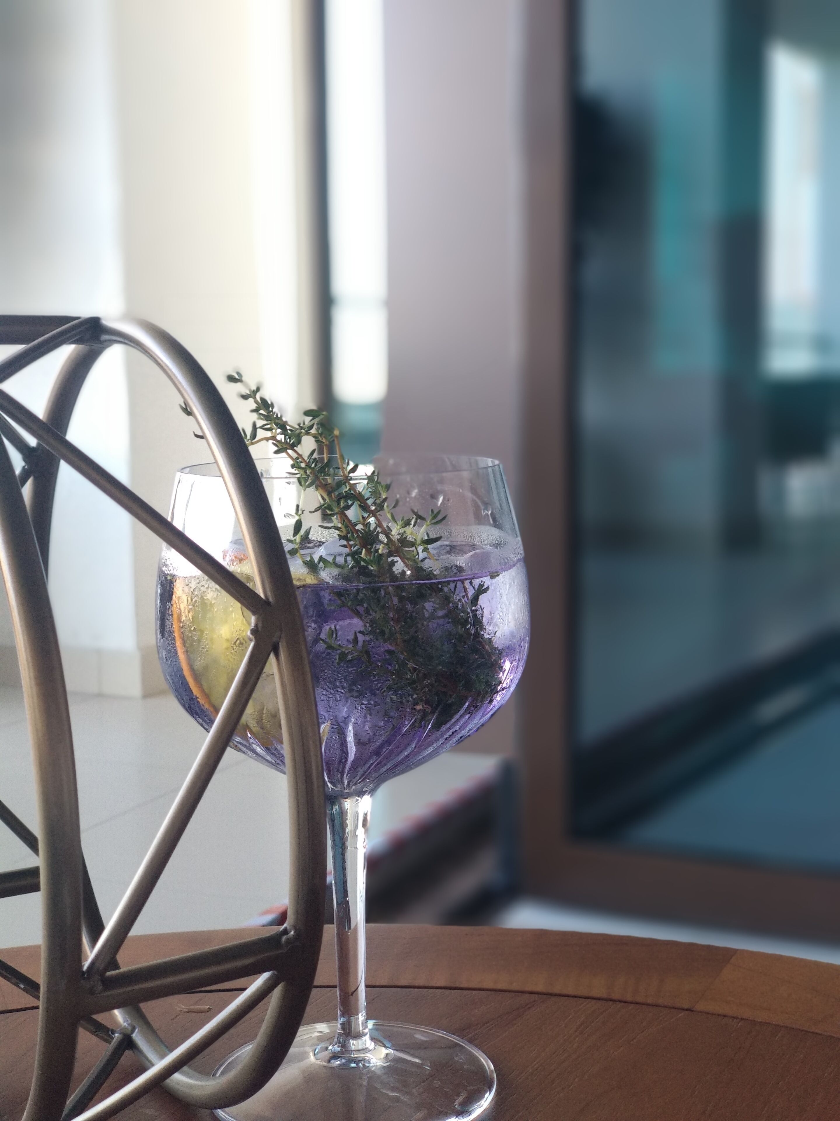 Un vaso con una bebida morada y un ramita de verdura, sobre una mesa cerca de una ventana.