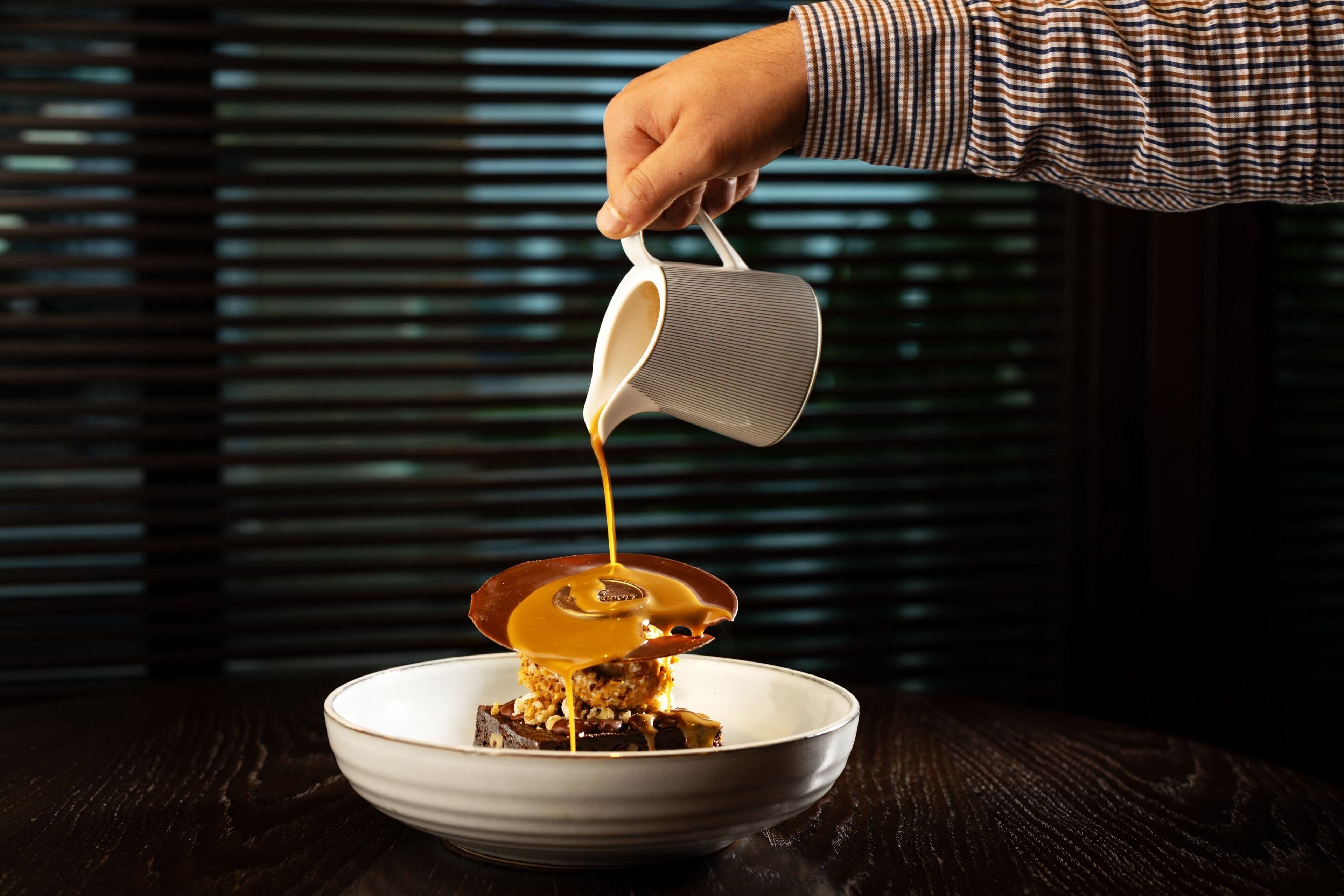 A hand pouring caramel sauce over a dessert on a white plate.