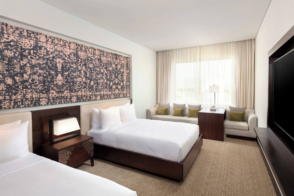 Habitación de hotel con una cama grande, una mesita de noche con lámpara, televisión, sofá y un amplio ventanal con cortinas.