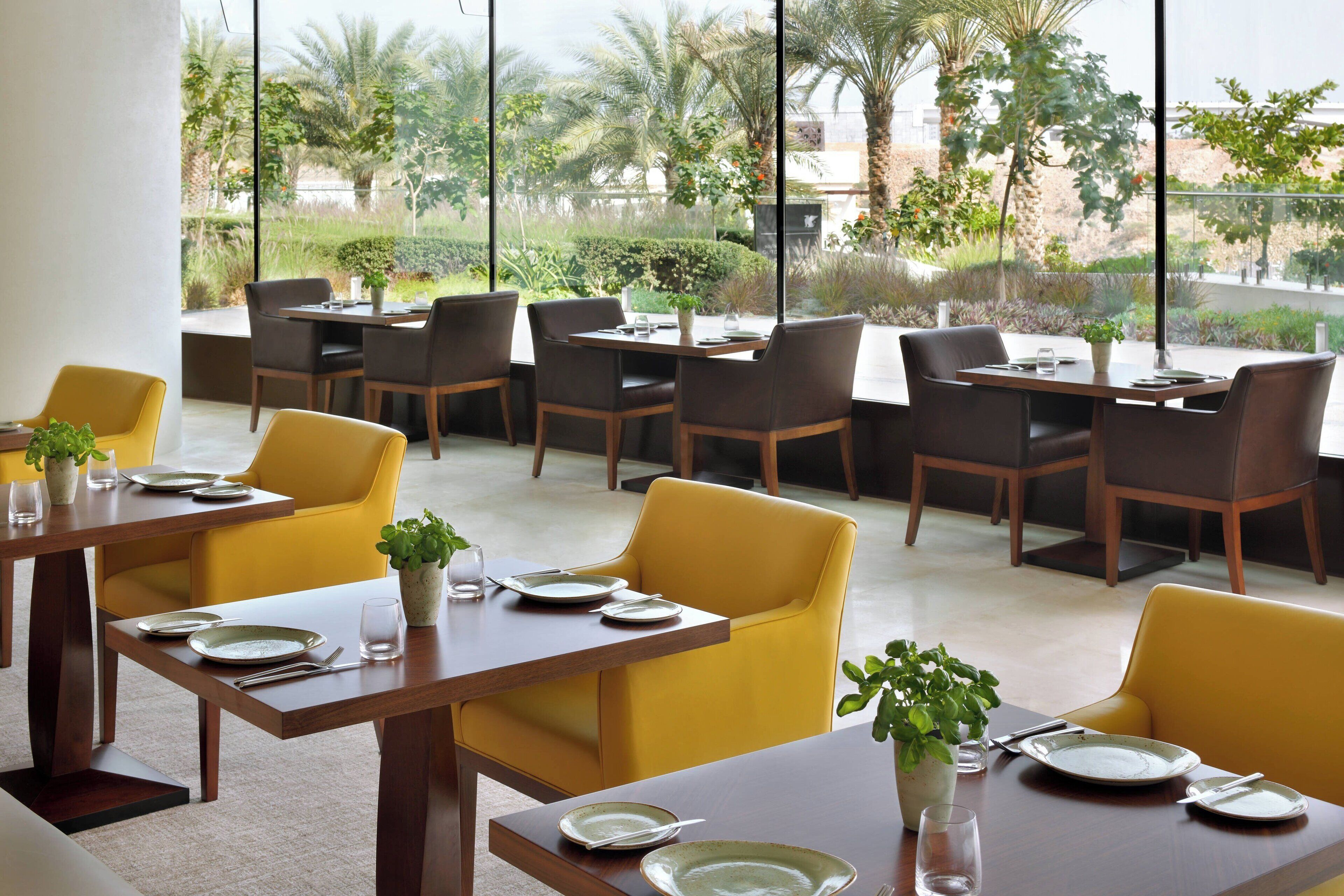 A dining area with yellow chairs and tables, large windows with a view of palm trees and greenery.