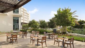 An outdoor seating area with multiple tables and chairs, surrounded by greenery and a clear sky.