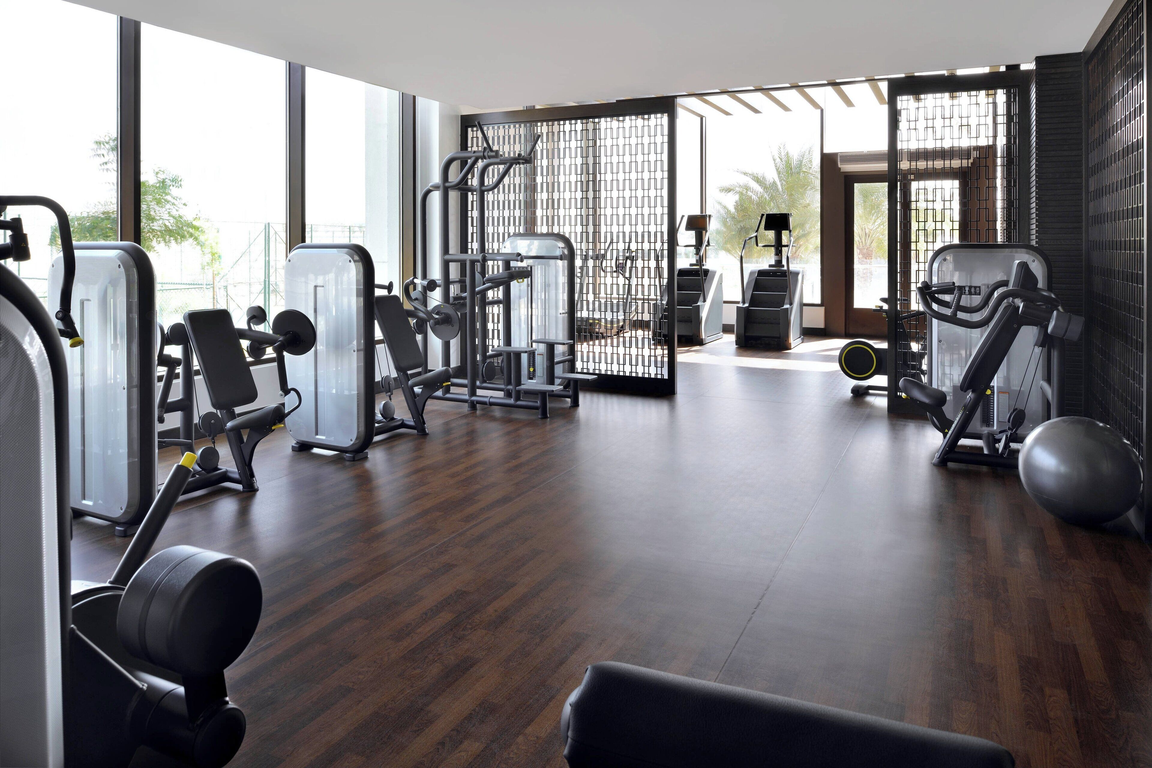 A modern gym with various exercise equipment, large windows, and a wooden floor.