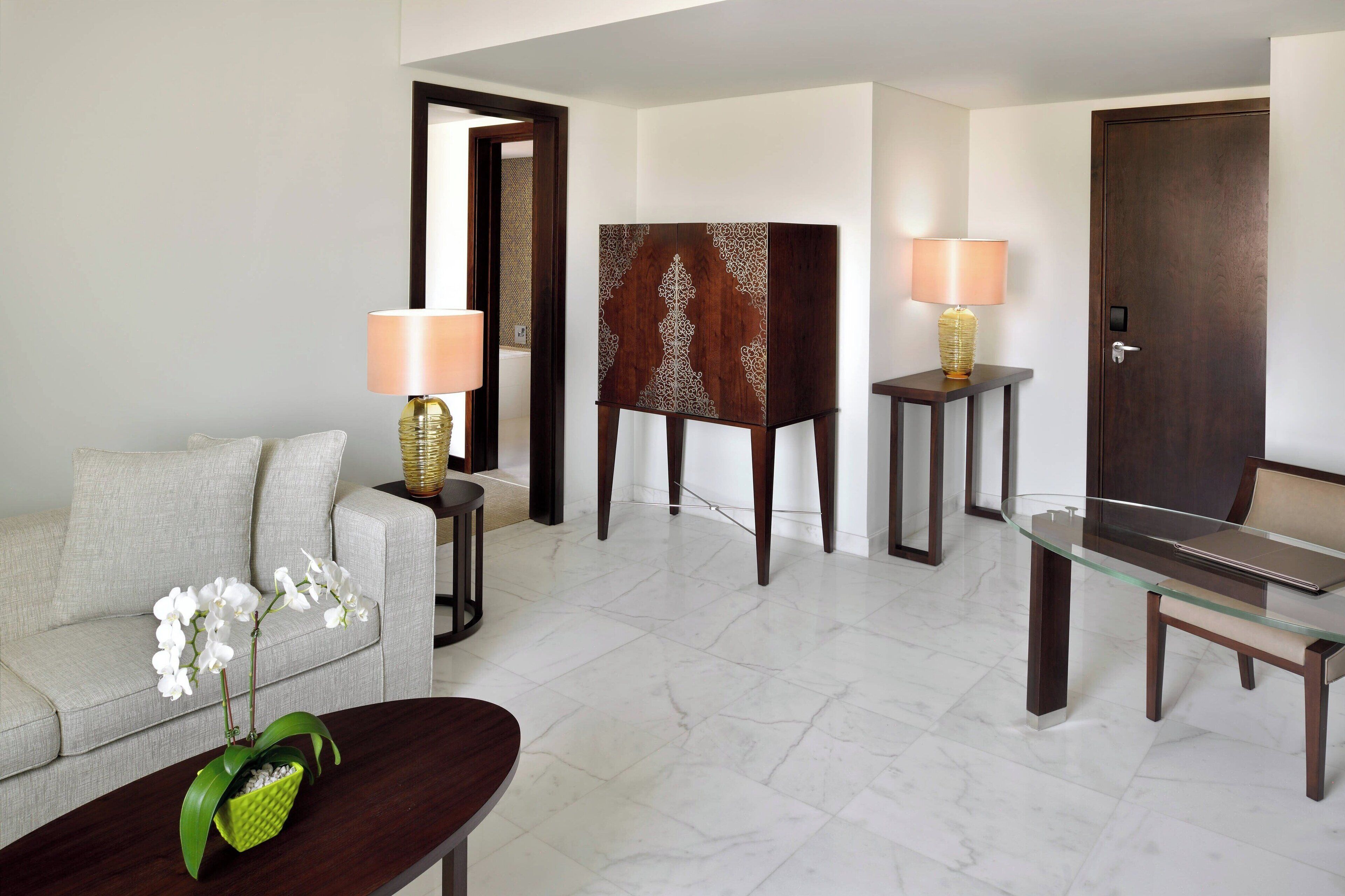 A modern living room with a sofa, a glass coffee table, two lamps, a decorative cabinet, and a door.