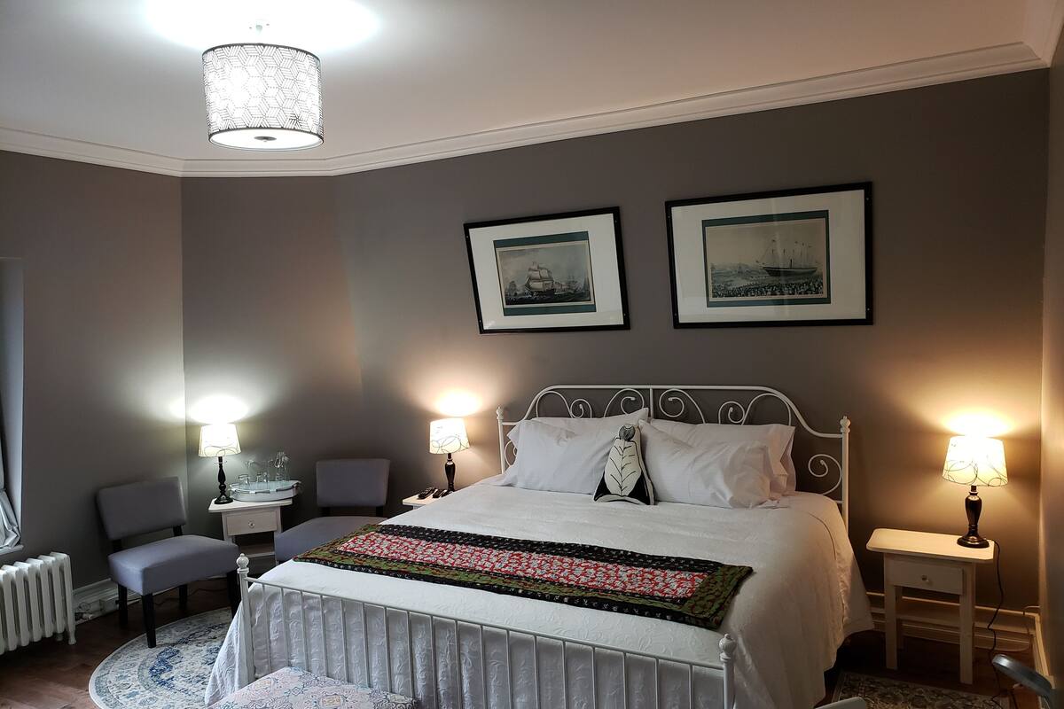 A bedroom with a bed, two bedside tables with lamps, a chair, a desk, and a radiator.