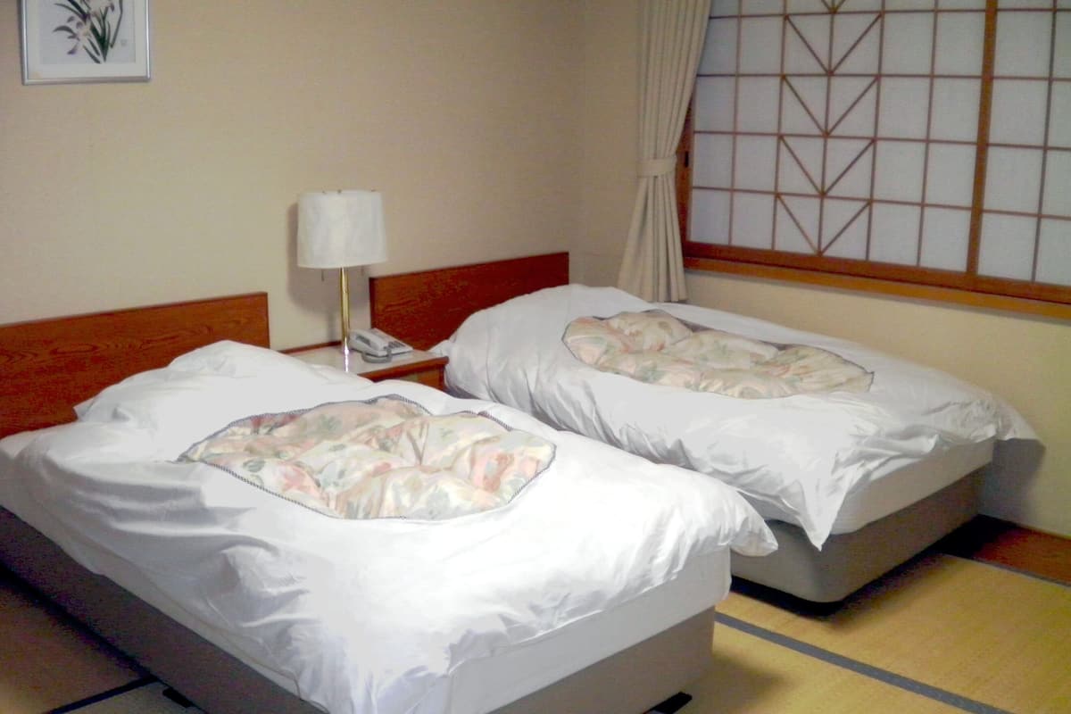 Dos camas individuales con ropa de cama floral, una mesita de noche con lámpara y una ventana con diseño de cuadrícula.