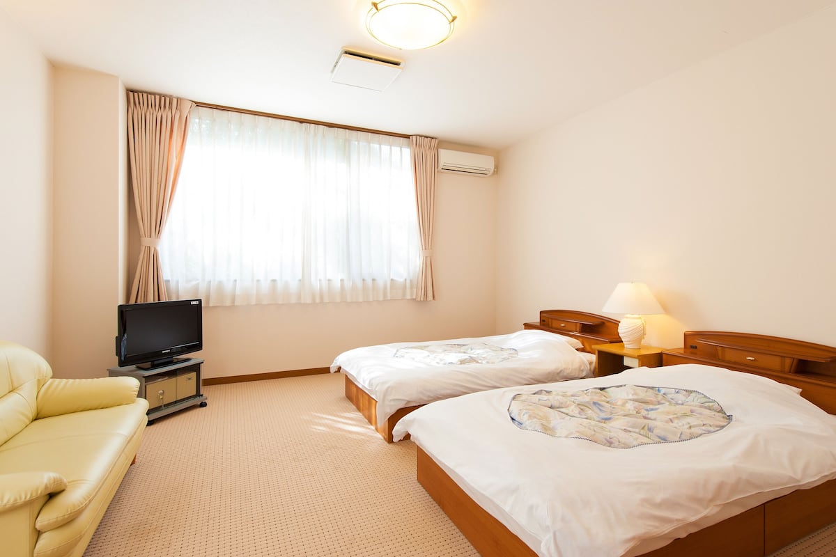 Habitación de hotel con dos camas, un televisor sobre un soporte, una lámpara y una ventana con cortinas.