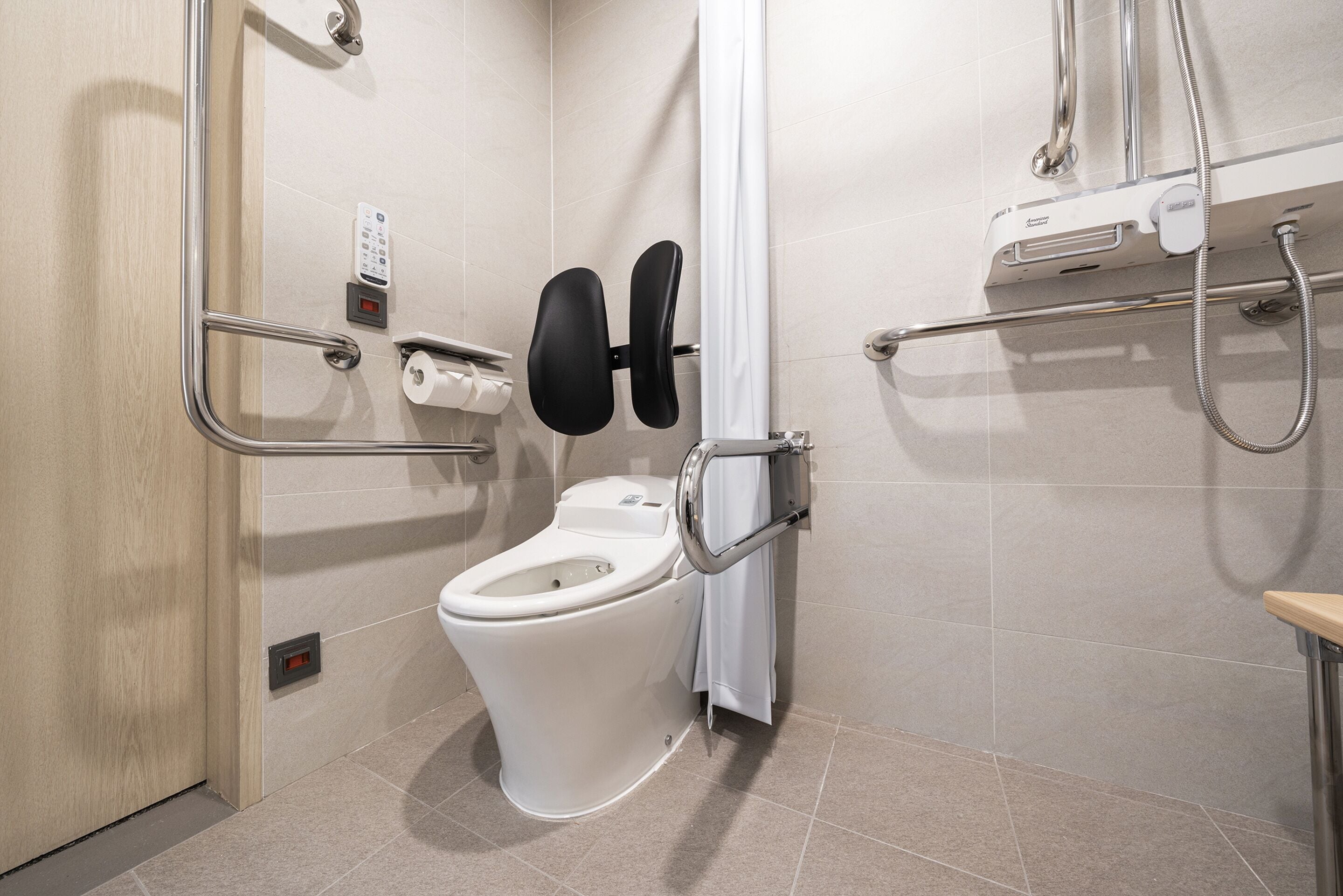 A modern bathroom with a toilet, grab bars, a shower control panel, and a hand dryer.