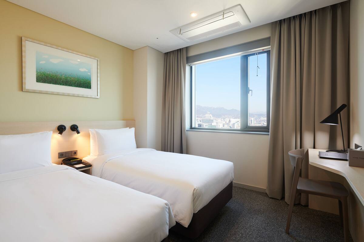 A hotel room with two beds, a desk, a chair, a large window with a view, and a painting on the wall.