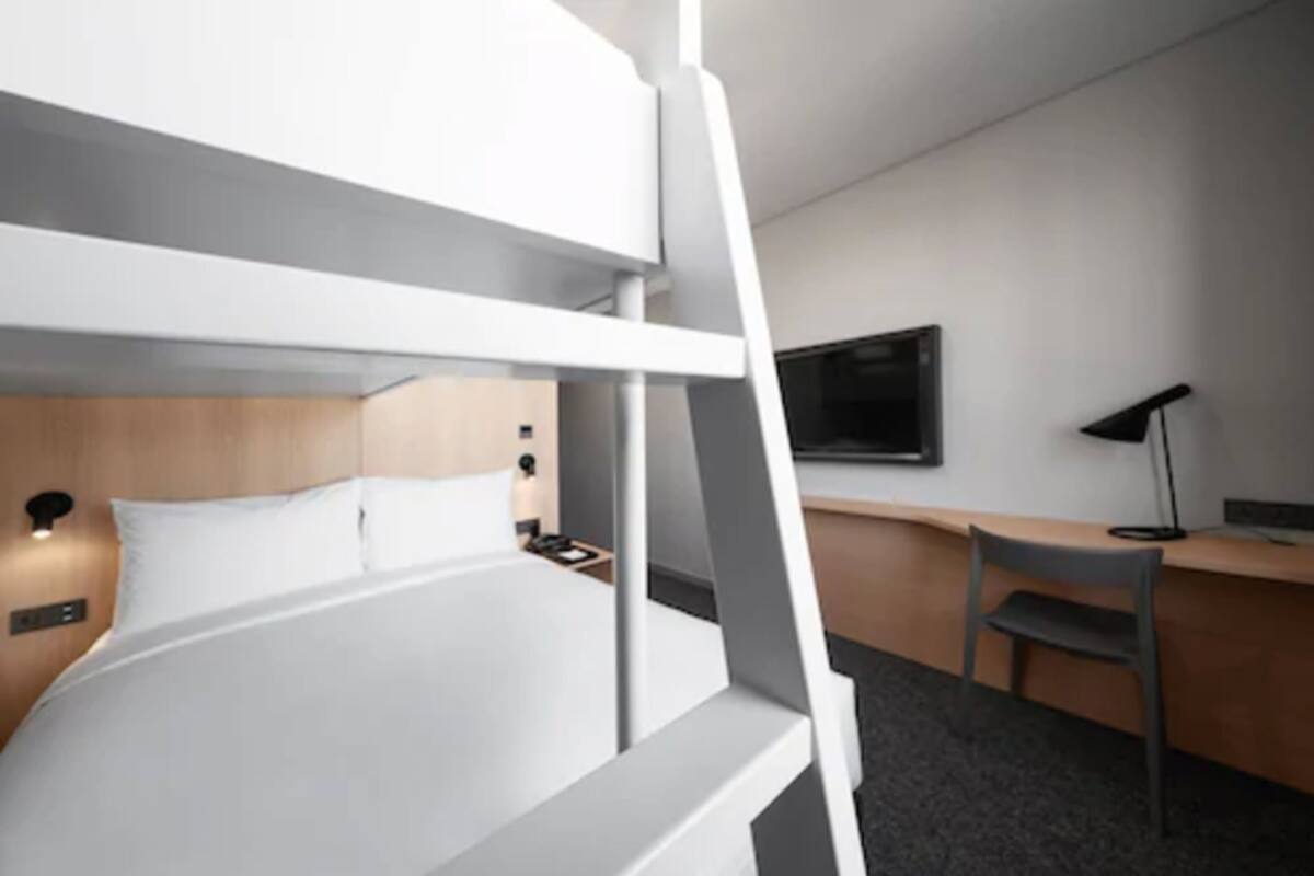 A modern hotel room with a bunk bed, a desk, a chair, a flat-screen TV, and a bedside lamp.