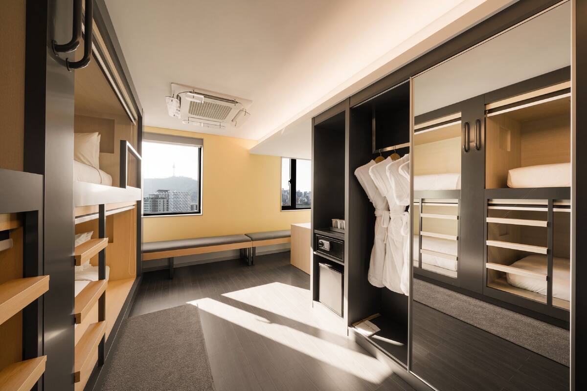 A modern hotel room with a bunk bed, a seating area, a wardrobe, and a window with a city view.