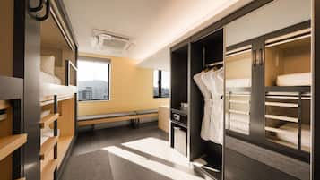 A modern hotel room with a bunk bed, a seating area, a wardrobe, and a window with a city view.