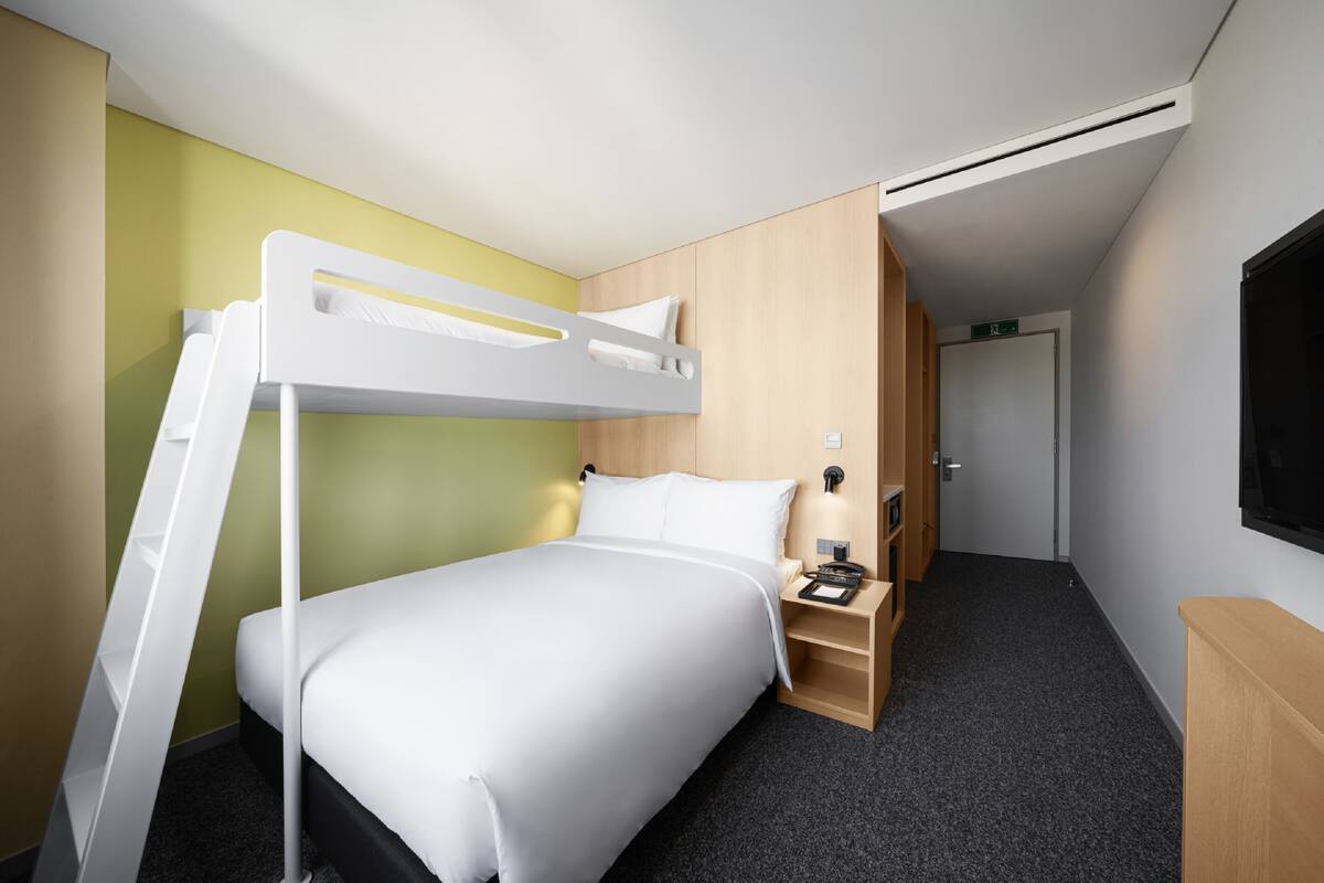 A modern hotel room with a bunk bed, a desk with a chair, a small shelf, and a TV mounted on the wall.