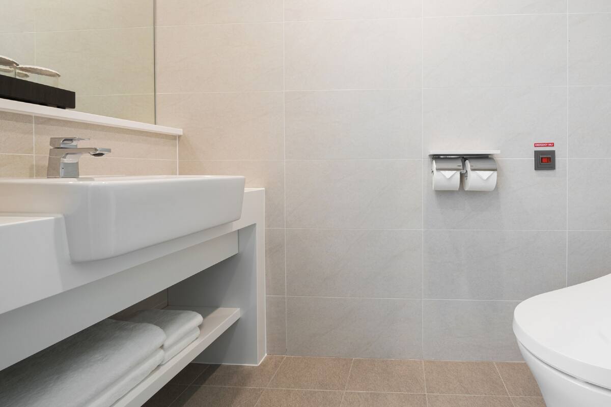 A modern bathroom with a white sink, mirror, and toilet. There are towels on a shelf and a toilet paper holder on the wall.