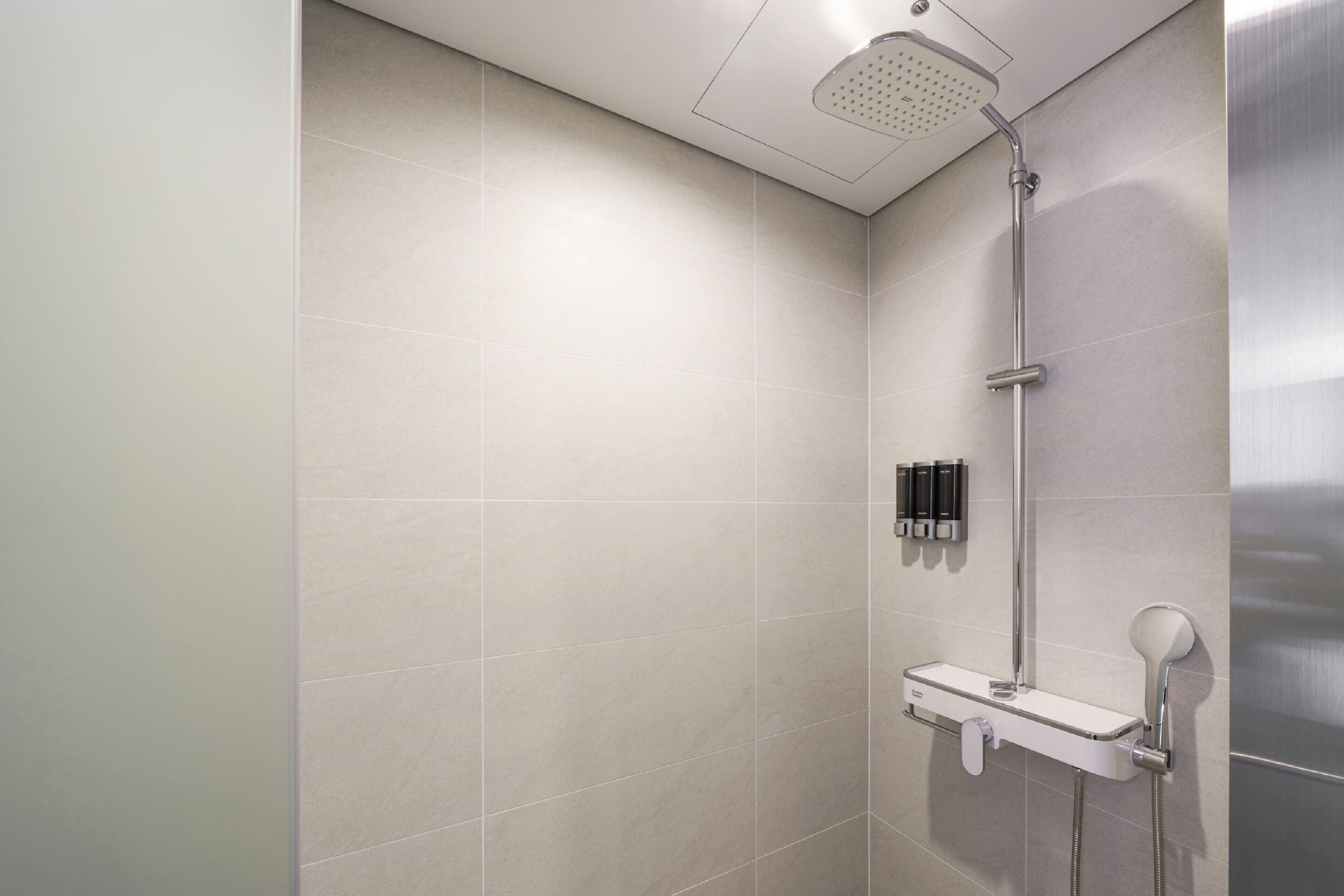 Una ducha moderna con cabezal de ducha manual, dispensador de jabón de pared y un estante pequeño.