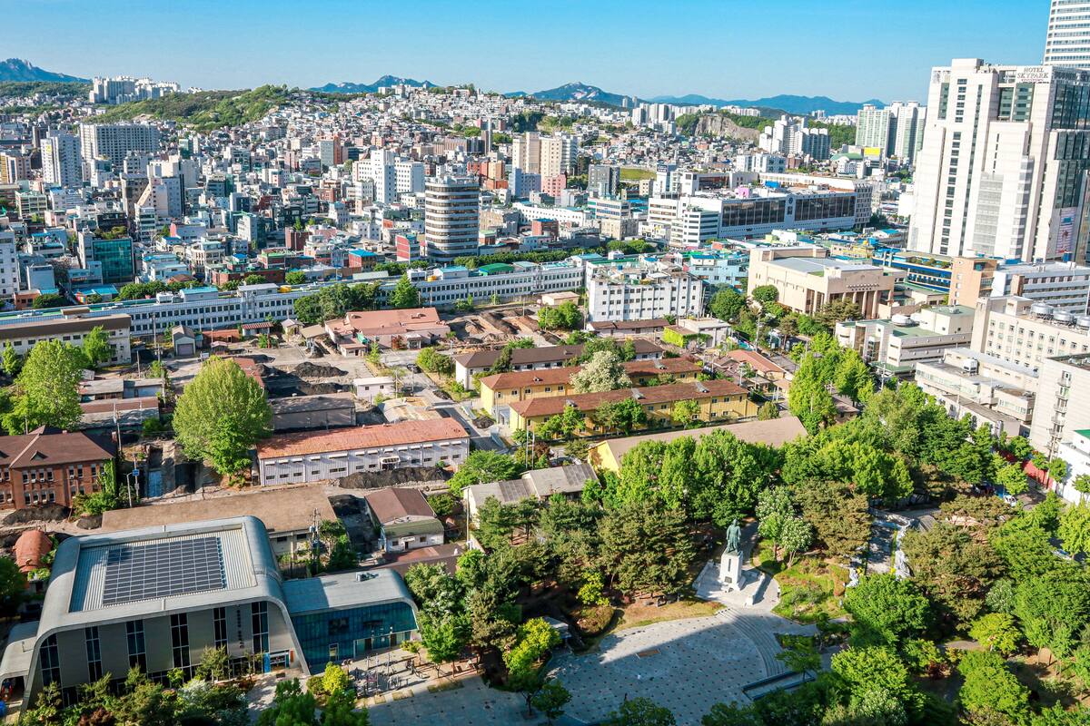 A cityscape with a mix of modern and older buildings, a central plaza with a statue, and a mountainous backdrop.