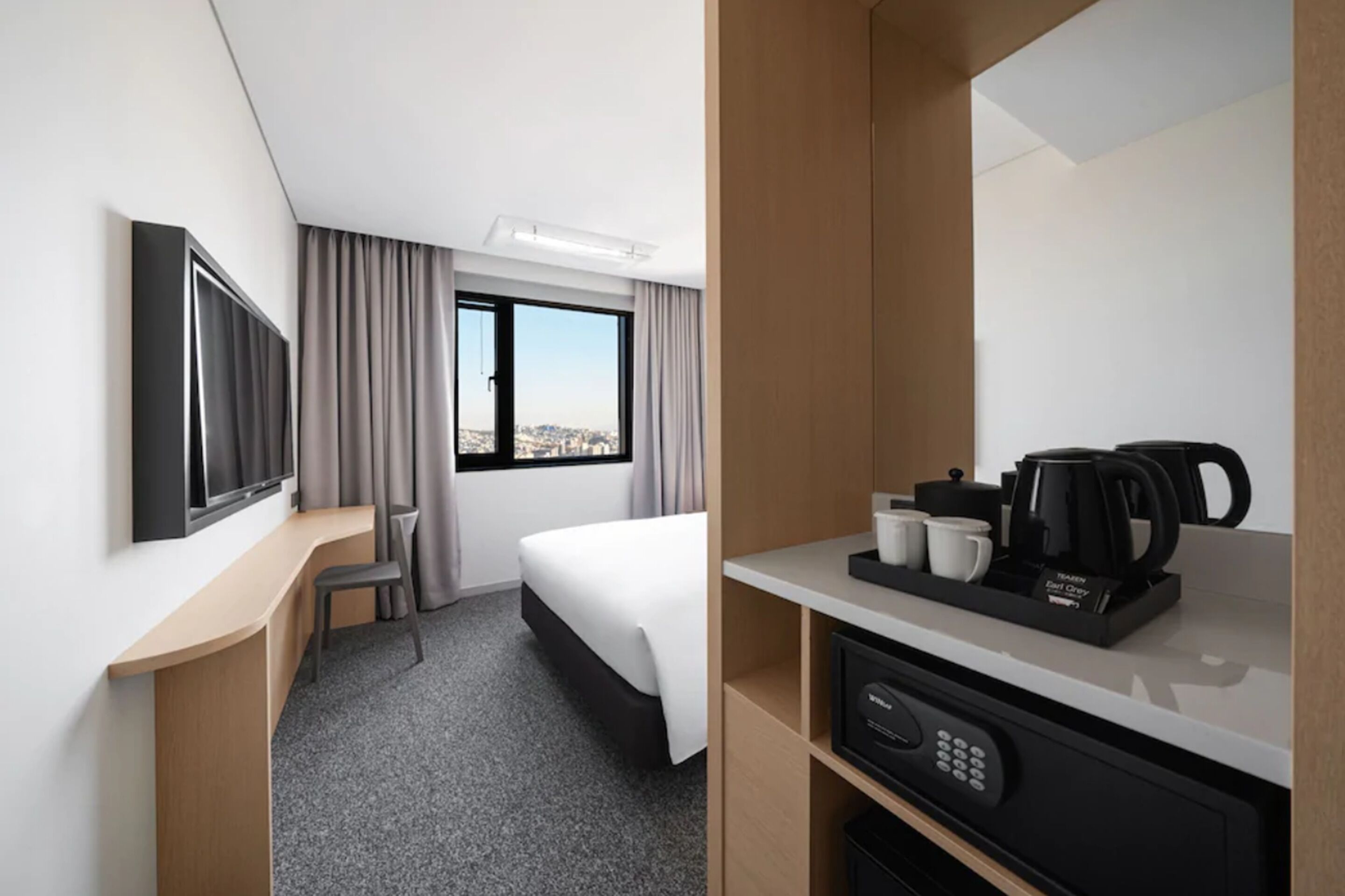 A hotel room with a bed, a desk with a coffee maker, a TV, and a window with curtains.