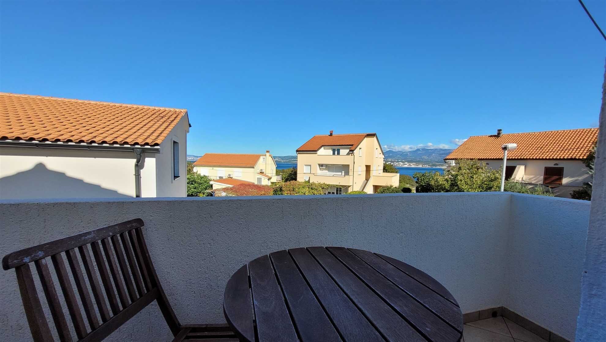 A balcony with a wooden table and chair, overlooking a coastal town with terracotta-roofed buildings and a clear blue sky.