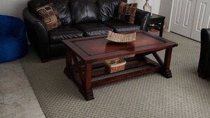 Un salón con un sofá de cuero negro, una mesa de centro de madera, un sillón de peluche azul, un cuadro enmarcado en la pared y una puerta cerrada al fondo.
