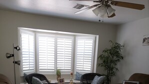 Una sala de estar con dos sofás, una mesa de centro y un ventilador de techo. Hay ventanas con persianas, una planta y un marco de fotos en el alféizar.
