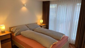 Un lit double avec un cadre en bois, deux tables de chevet avec des lampes, une fenêtre avec des rideaux légers et un plancher en bois.